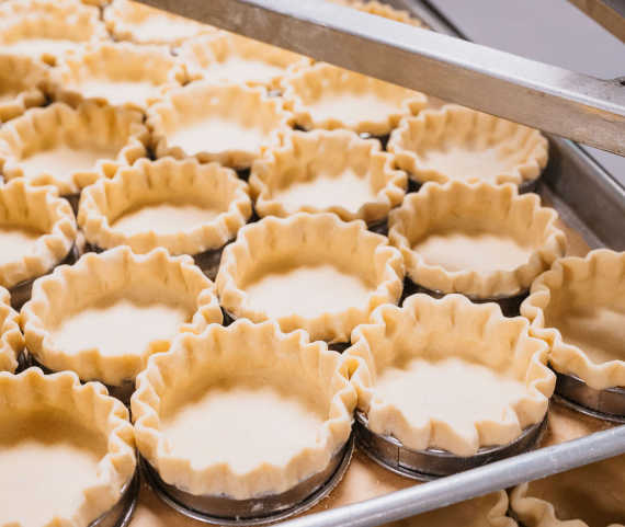 Unbaked tart shells filled with white filling in a bakery. A metal ruler is visible at the top of the tray.