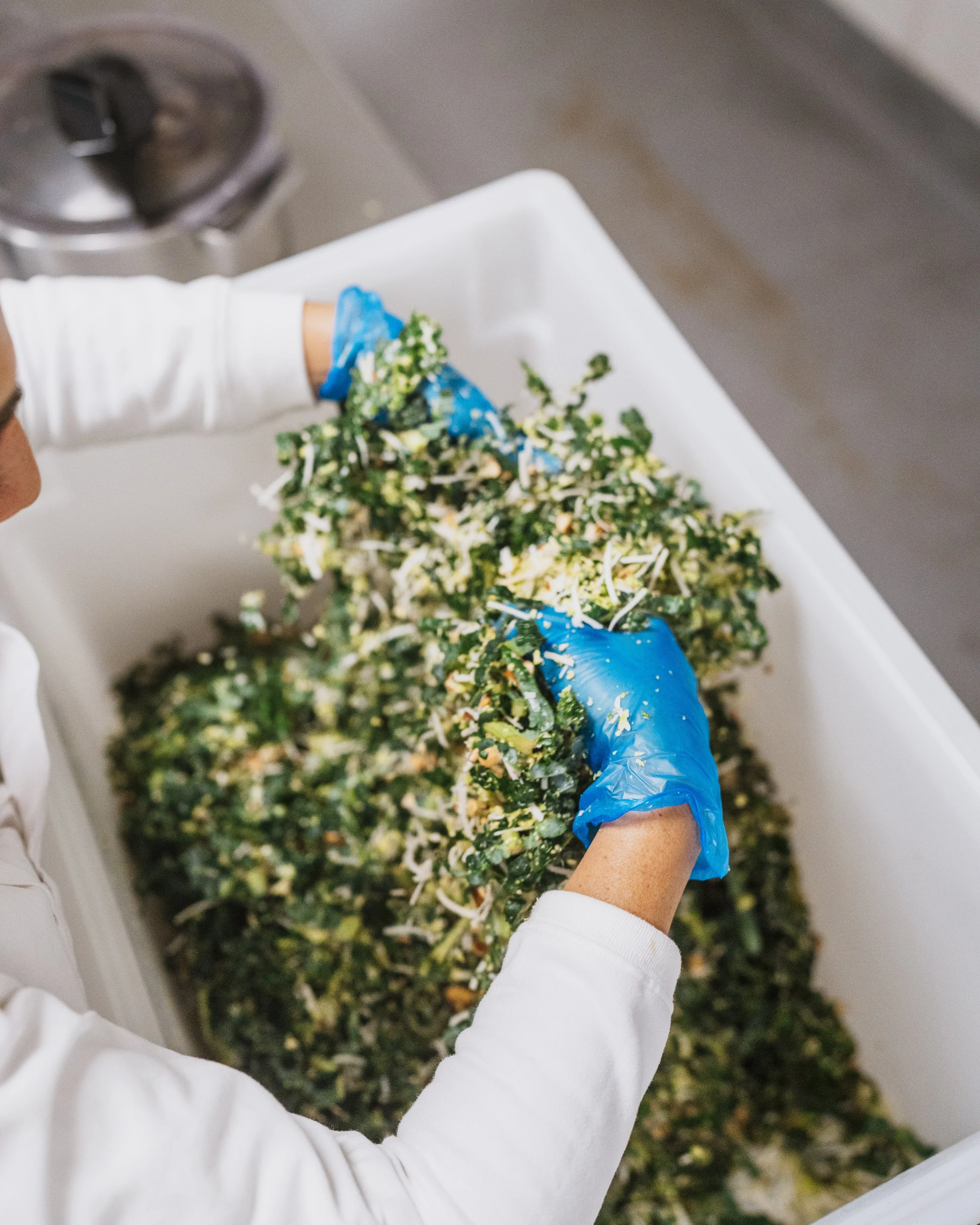 A person wearing a white long-sleeve shirt and blue gloves handling shredded paper or confetti over a large white container or bin, with an industrial machine and some green ground in the background.