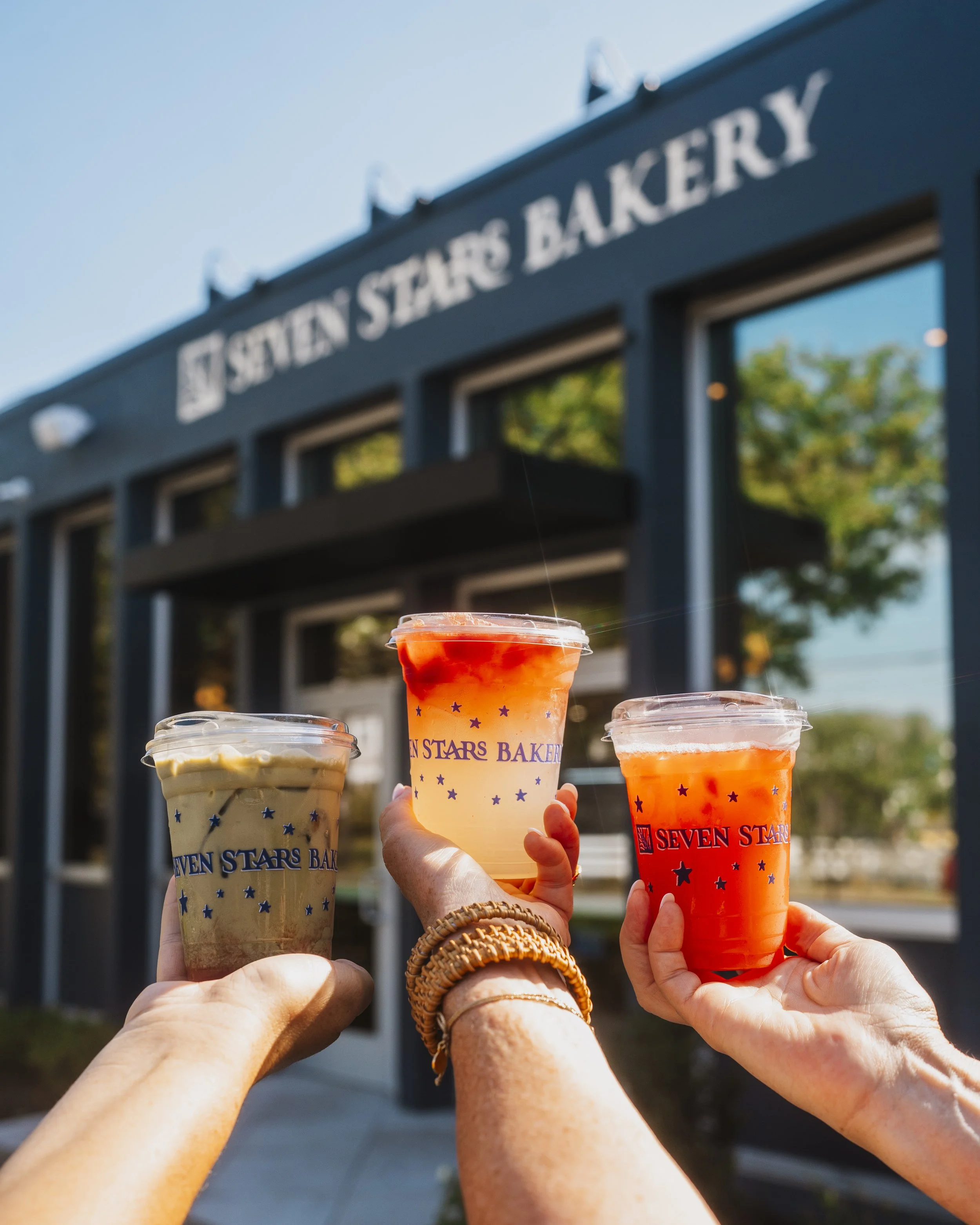 Friends toasting with Seven Stars Bakery iced drinks outside the Edgewood Cranston café on a sunny day.