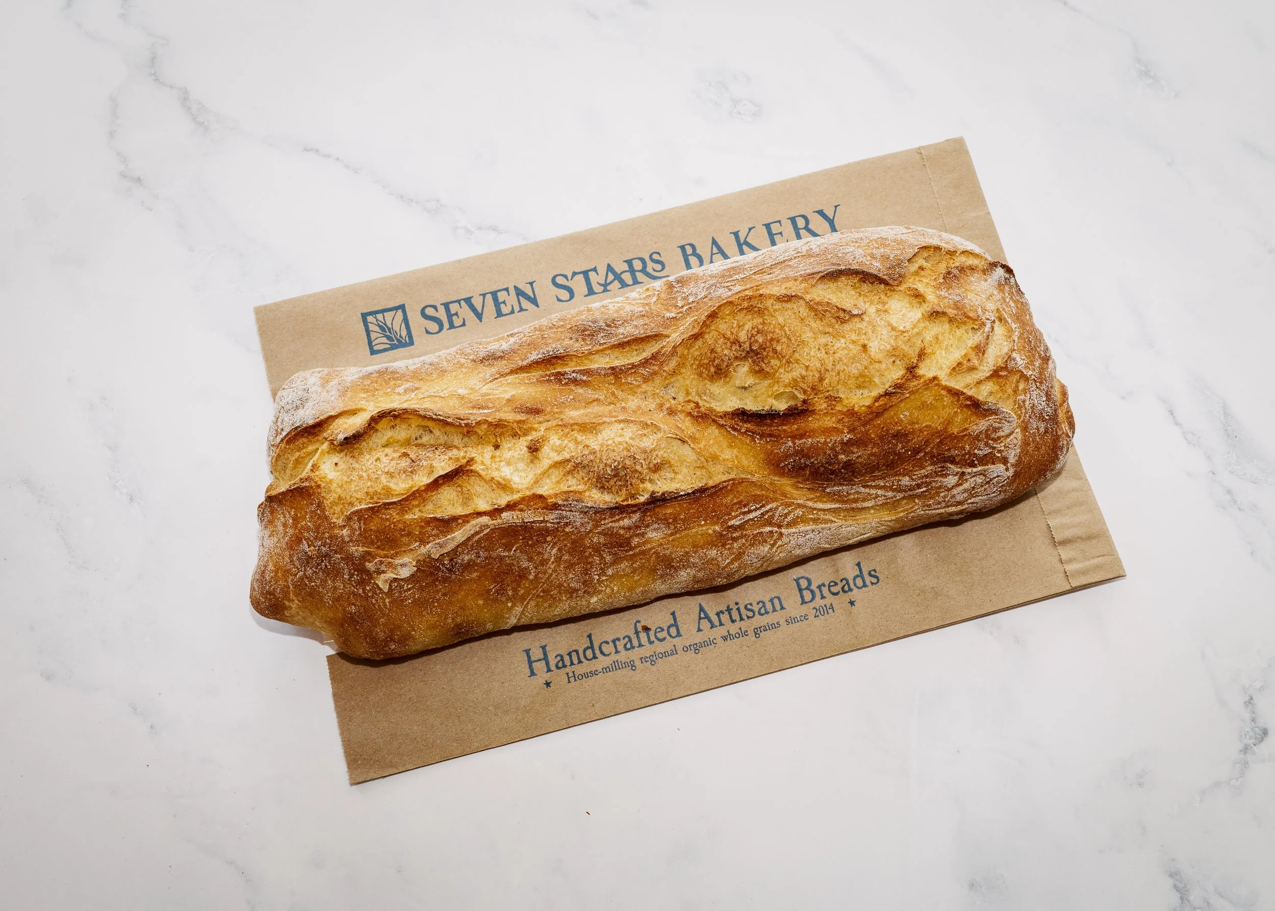 A loaf of durum with a golden-brown crust resting on a brown paper bag from Seven Stars Bakery, on a white marble surface.
