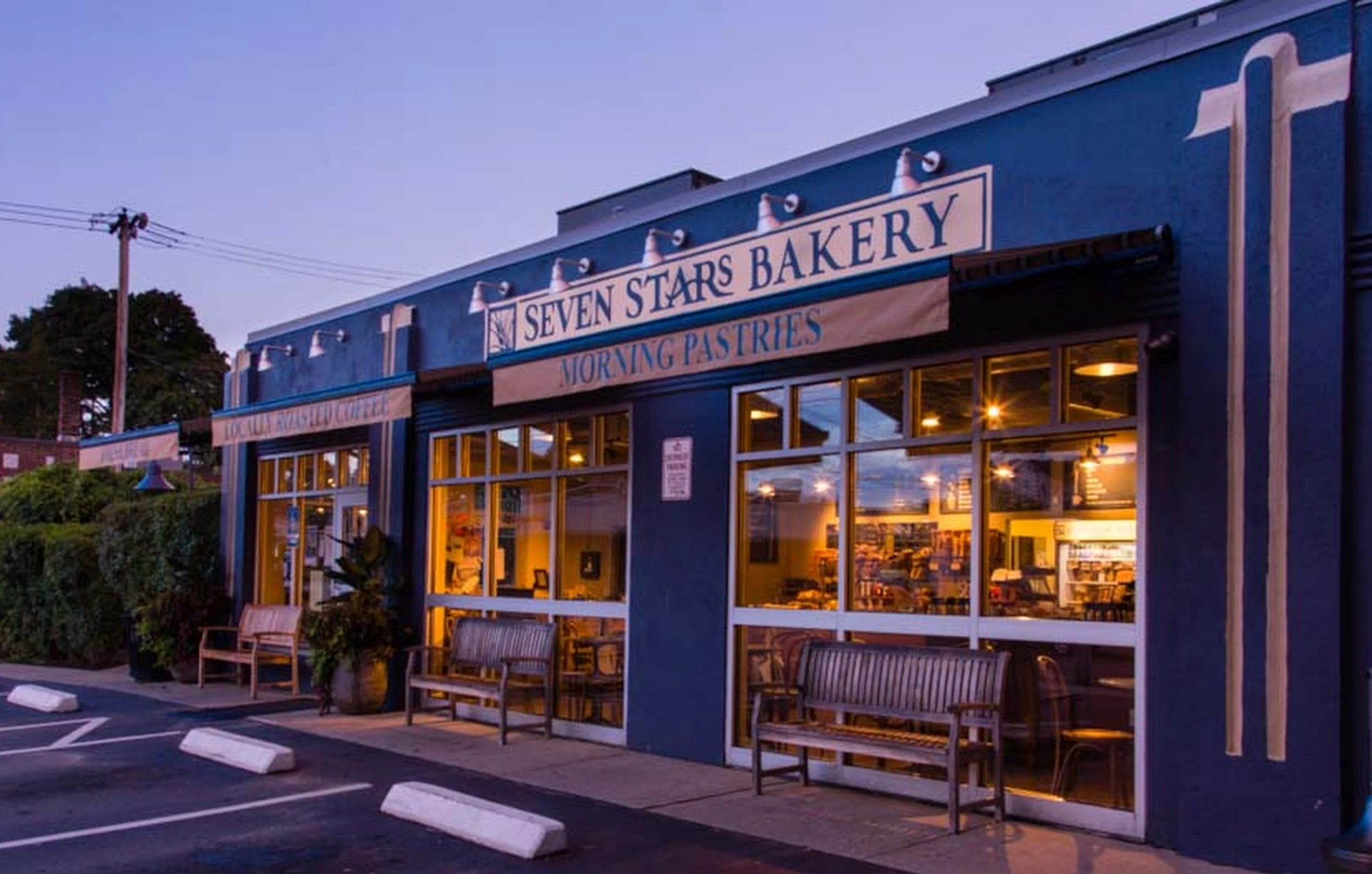 The exterior of Seven Stars Bakery during dusk, showing a blue building with large glass windows, interior lighting, and signage indicating Morning Pastries; there are outdoor benches, a parking lot with white parking blocks, and a utility pole with power lines nearby.