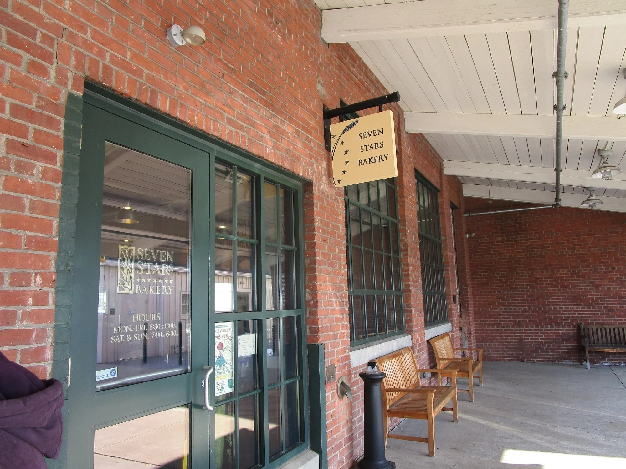 Brick storefront and hanging sign of Seven Stars Bakery Rumford, with benches out front.