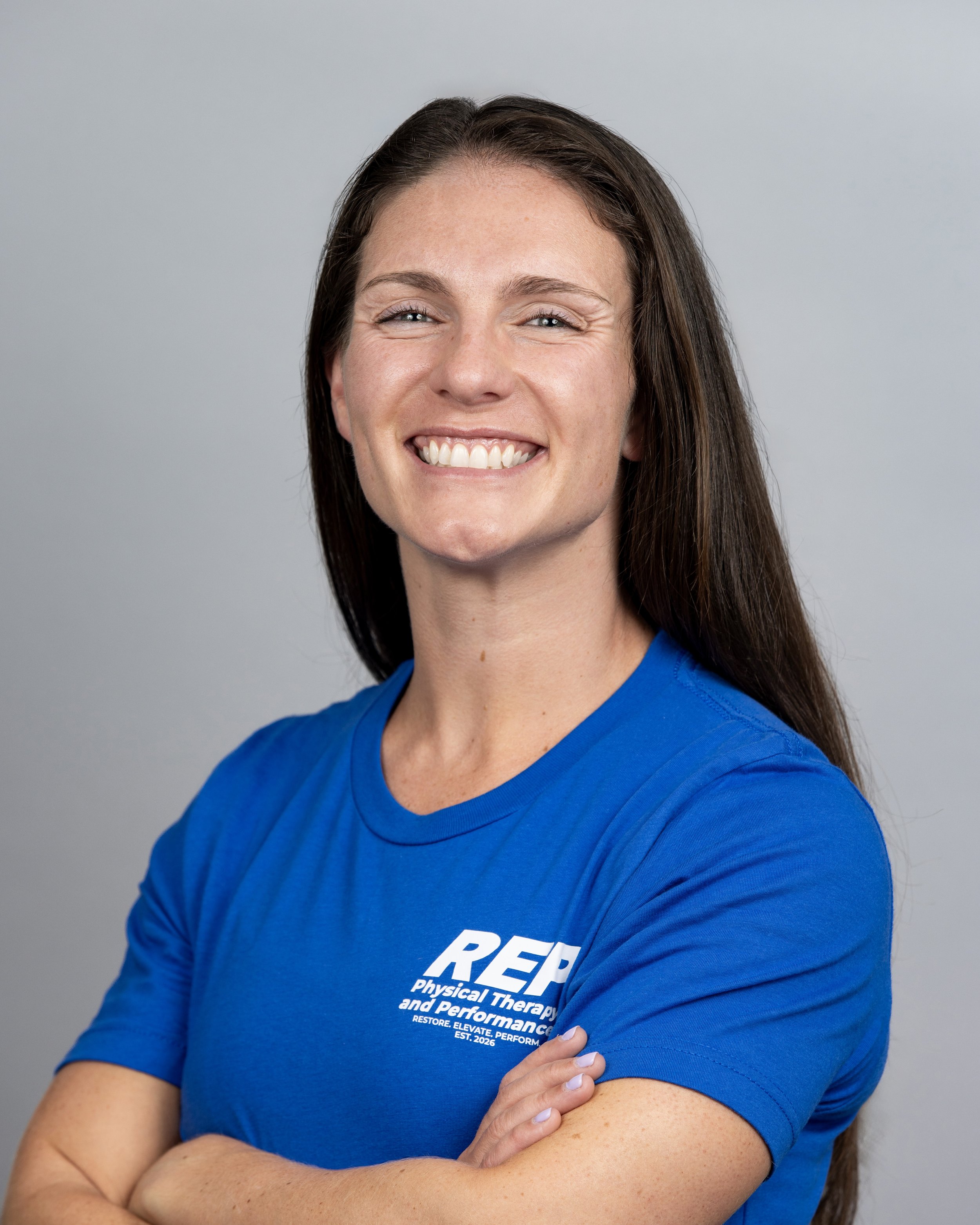 Dr. Ciara Forde, PT, DPT, CSCS, a physical therapist in Melbourne, Florida. A woman with long brown hair wearing a blue t-shirt with white text, smiling and crossing her arms against a plain light gray background.