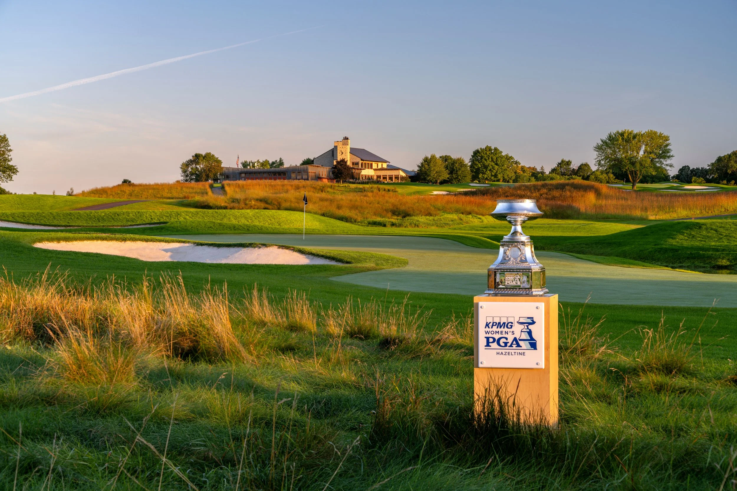 A golf course with a putting green, sand trap, and flag, near a clubhouse. A trophy is on a pedestal in the foreground with a sign reading 'KPMG Women's PGA, Hazeltine.'