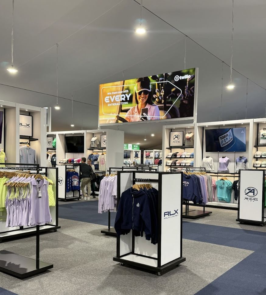 Inside a retail store selling athletic apparel and accessories, with clothing racks displaying shirts, hoodies, and jackets, and digital advertising screens overhead.