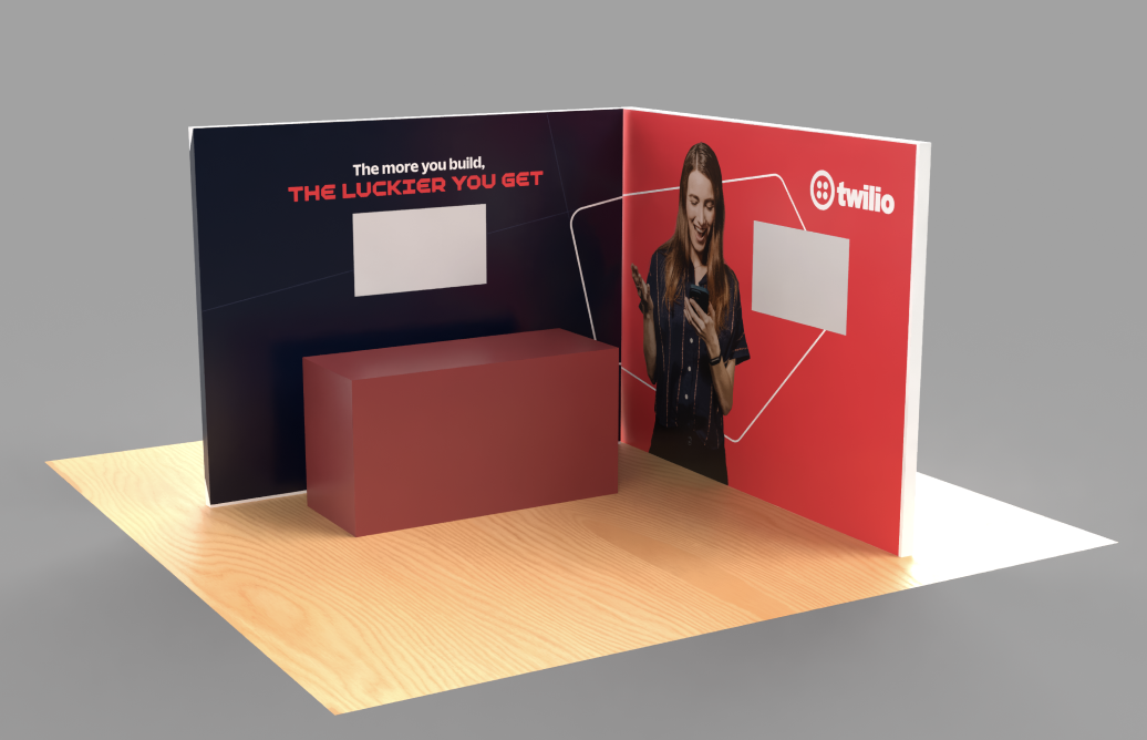 A trade show booth for Twilio with a curved backdrop, featuring a woman on the right side looking at her phone, and a rectangular red table in front. The backdrop has text that says "The more you build, the luckier you get" and Twilio's logo.