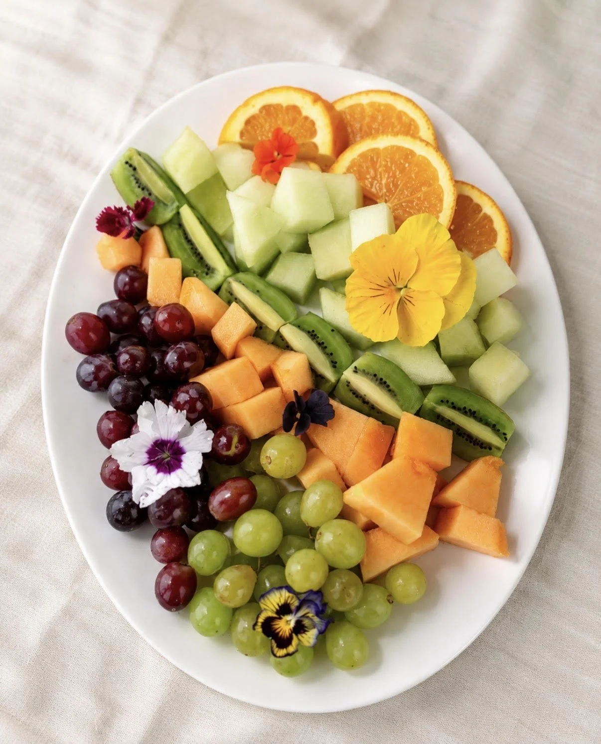 Assorted fresh fruit including orange slices, honeydew melon, kiwi slices, cantaloupe, green and red grapes, with colorful edible flowers on a white oval plate.