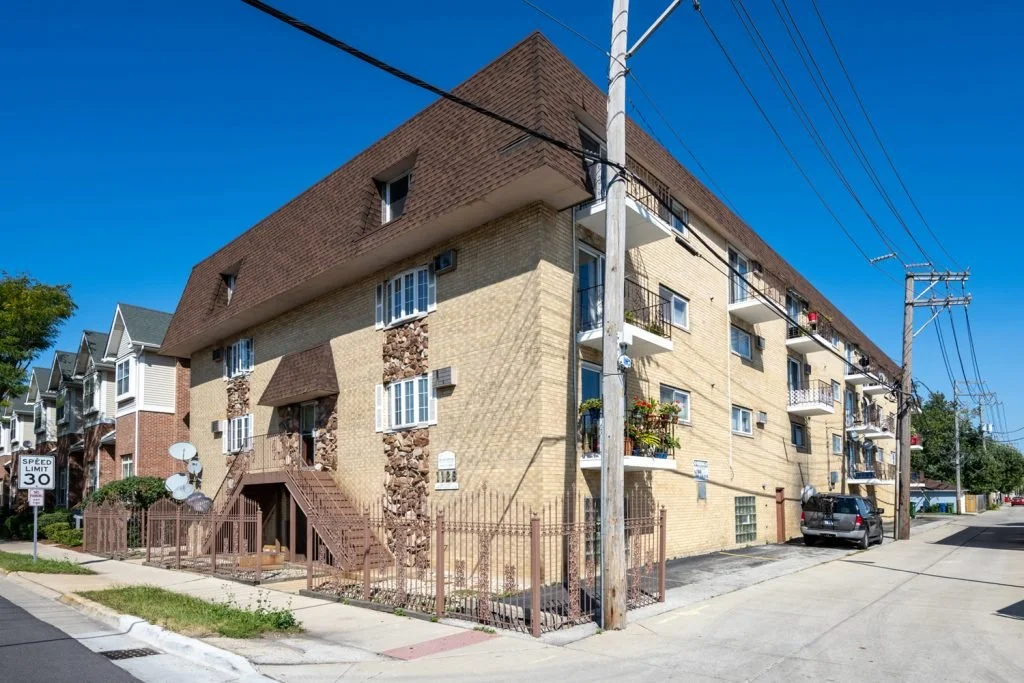 Multi-story residential building with beige brick exterior, balconies, and satellite dishes, located on a street corner with utility poles and a 30 mph speed limit sign.
