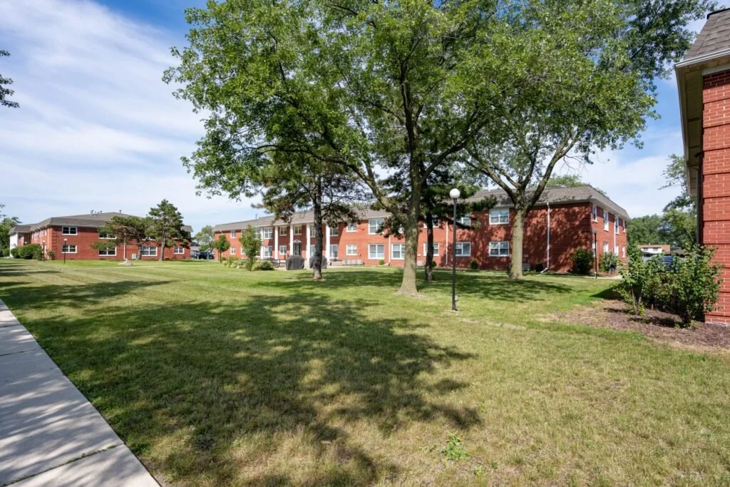 A grassy courtyard with trees, brick apartment buildings, a sidewalk, and a lamppost, under a blue sky with light clouds.