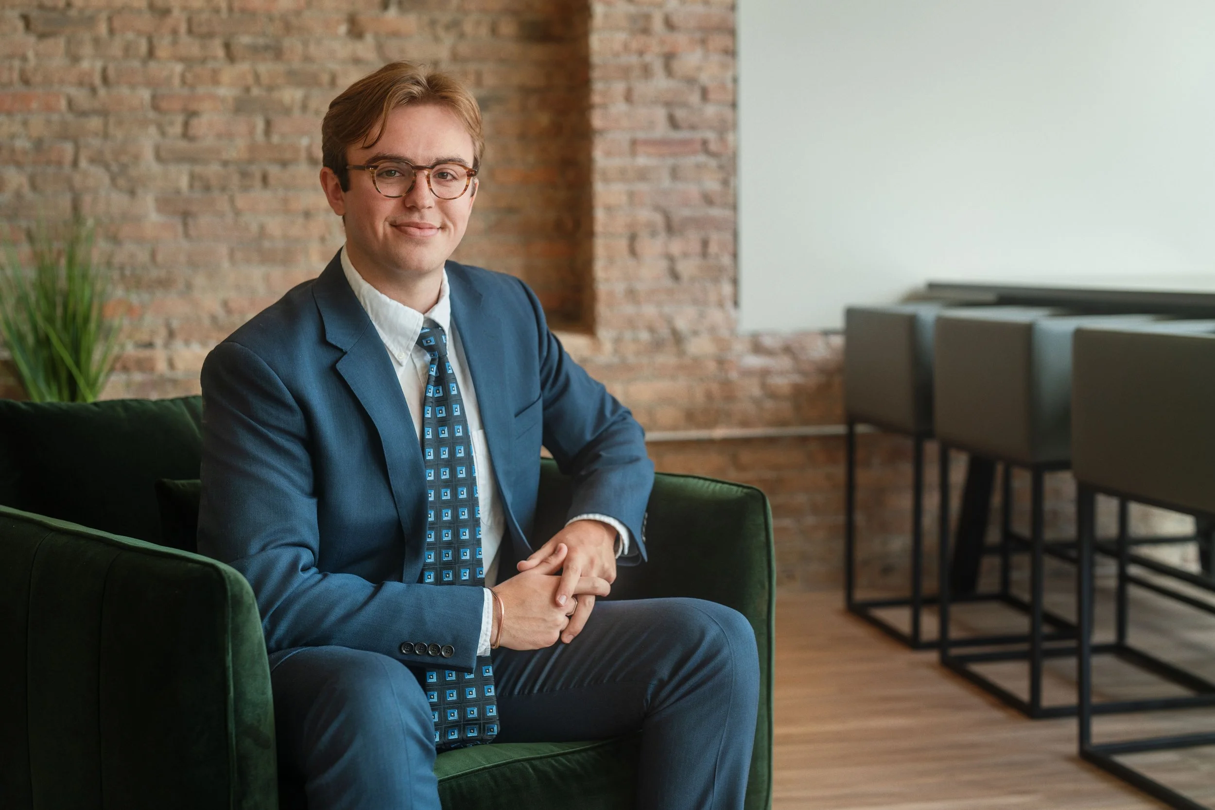 A young man with glasses and light brown hair, wearing a blue suit and a patterned tie, is sitting on a green chair in a modern room with a brick wall on one side and white walls on the other. He has a slight smile and is looking at the camera.