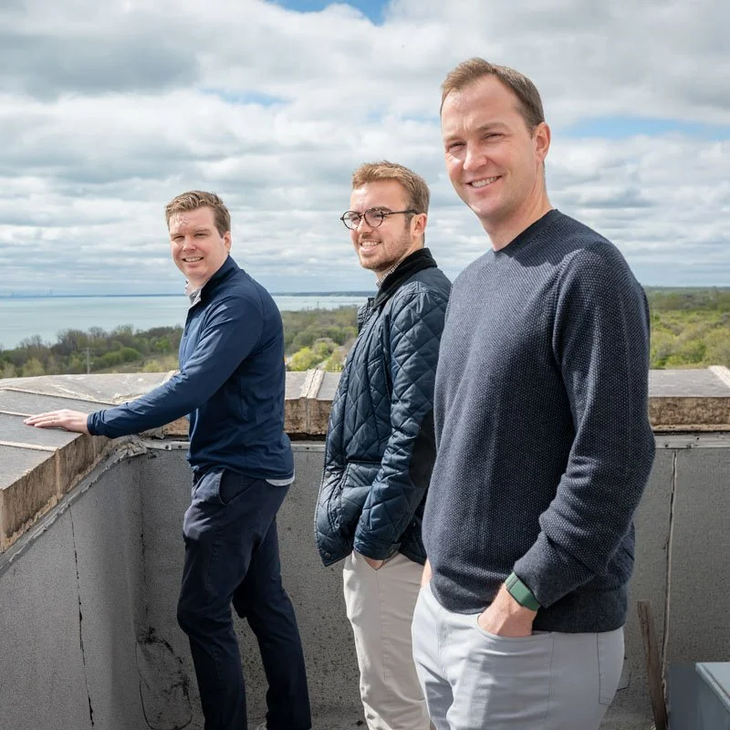 Three men standing on a rooftop with a scenic view of trees, water, and a cloudy sky in the background.