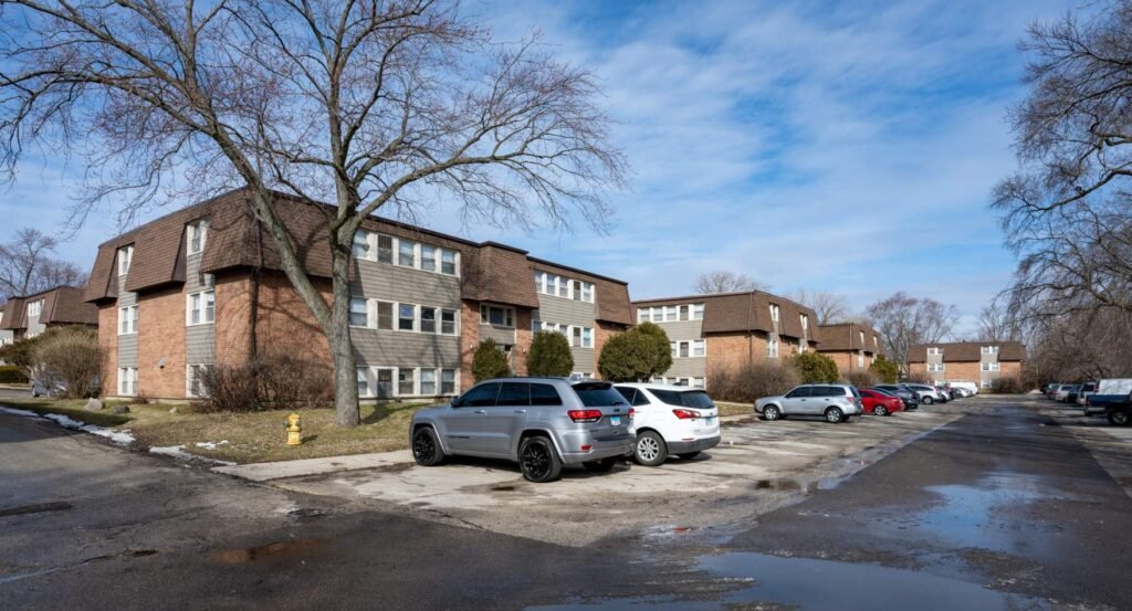 A residential apartment complex with brick buildings, leafless trees, and a parking lot with several parked cars on a clear day.