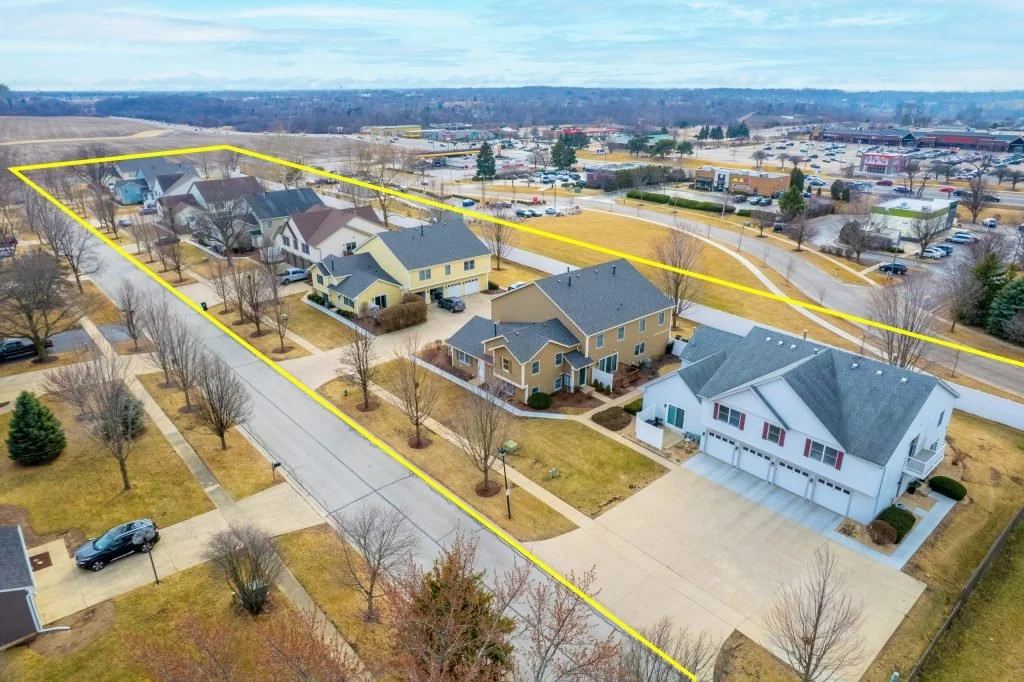 An aerial view of a residential neighborhood with several houses, trees, and a large property boundary outlined in yellow, nearby commercial area with parking lot and stores.