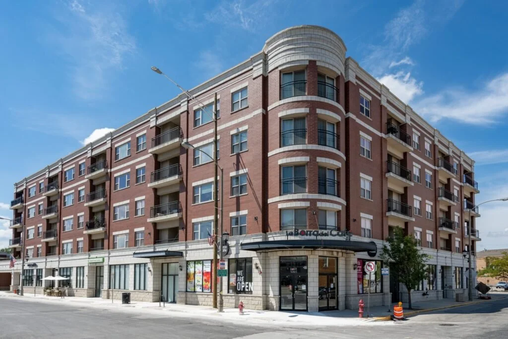 A multi-story brick and concrete building with retail shops on the ground floor and residential apartments above, located on a corner street with a blue sky background.