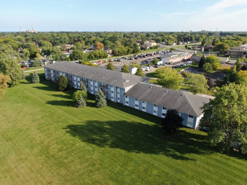 An aerial view of a multi-story apartment complex surrounded by green lawns and trees, with a parking lot and additional buildings in the background.