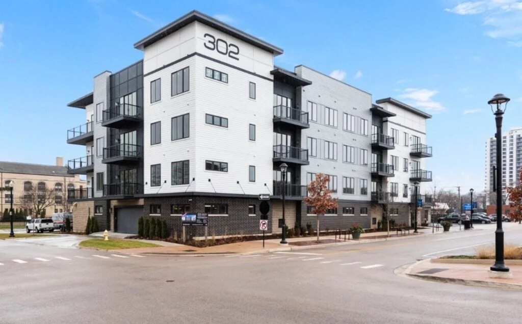 Modern four-story apartment building with white exterior, black balconies, and the number 302 displayed on the top corner, located at a street corner with street lamps and small trees.