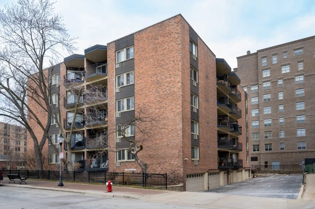 A mid-rise brick apartment building with multiple balconies, leafless trees in front, and a parking garage at the ground level, with a sidewalk and street lamp in the foreground.