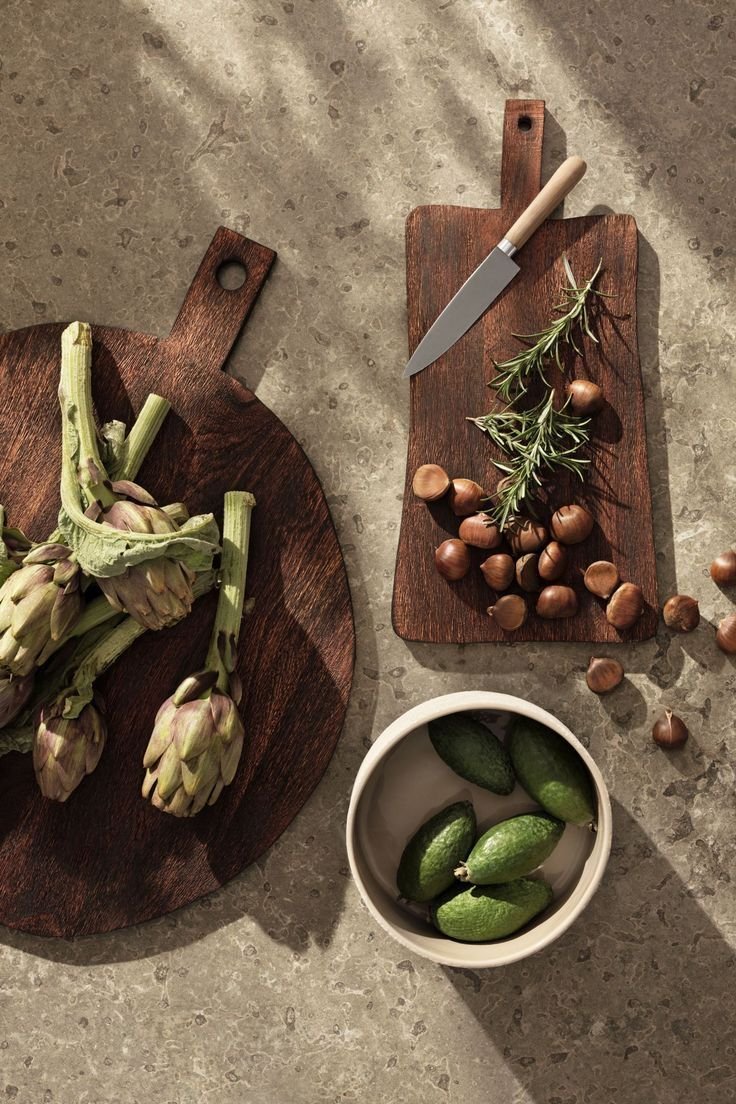 Fresh artichokes, hazelnuts, cucumbers, garlic, and rosemary with a knife on two cutting boards and a white bowl placed on a gray countertop.