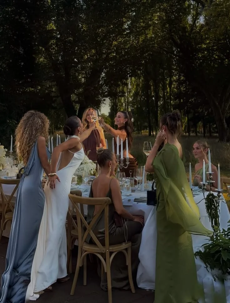 A group of women celebrating at an outdoor dinner party during sunset, raising glasses in a toast, with a long table set with candles and floral arrangements amidst trees.