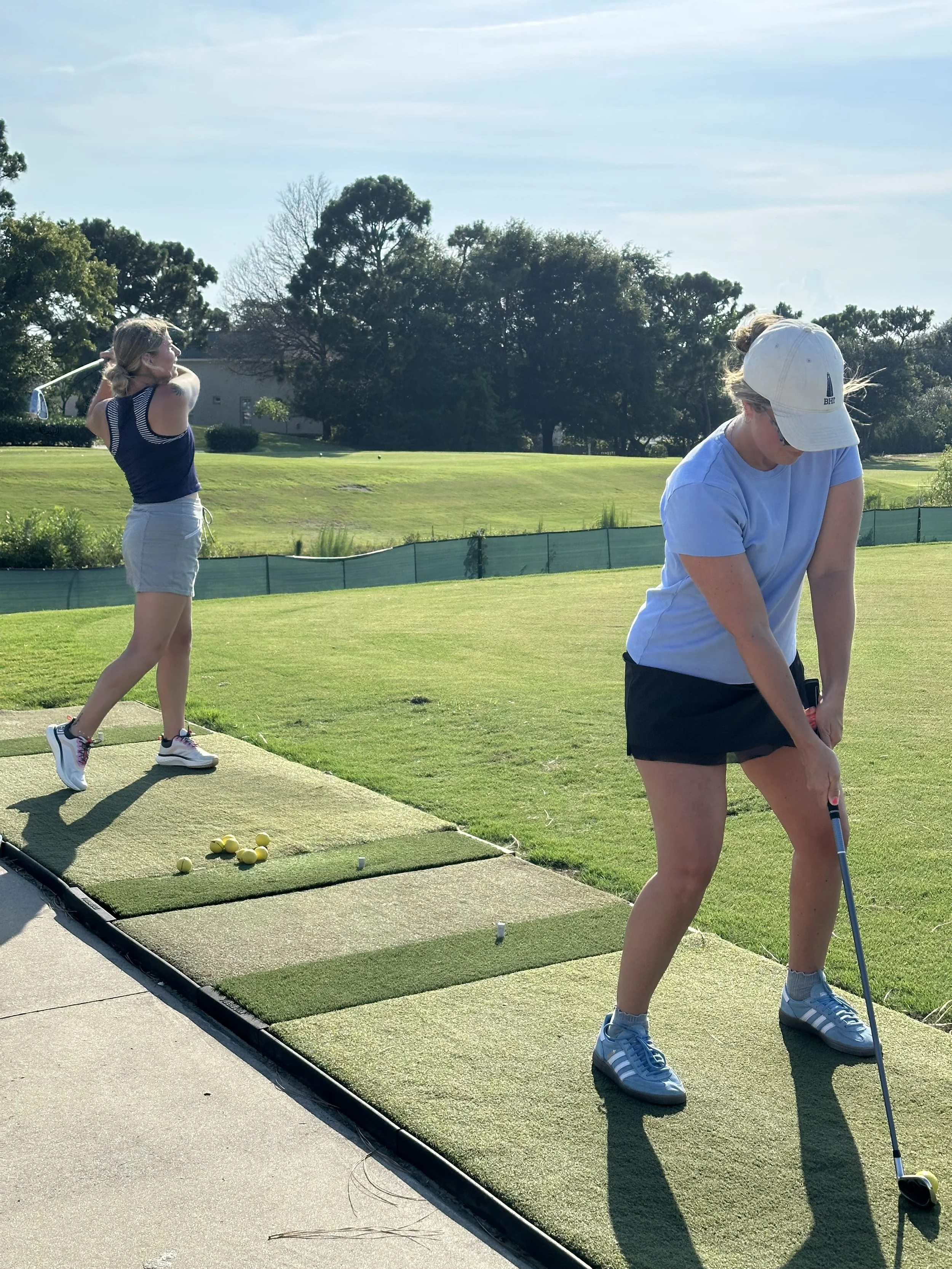 Two women practicing golf at a driving range. One woman is swinging a golf club, while the other is preparing to hit the ball. There are golf balls on the mat and a golf club in her hand.