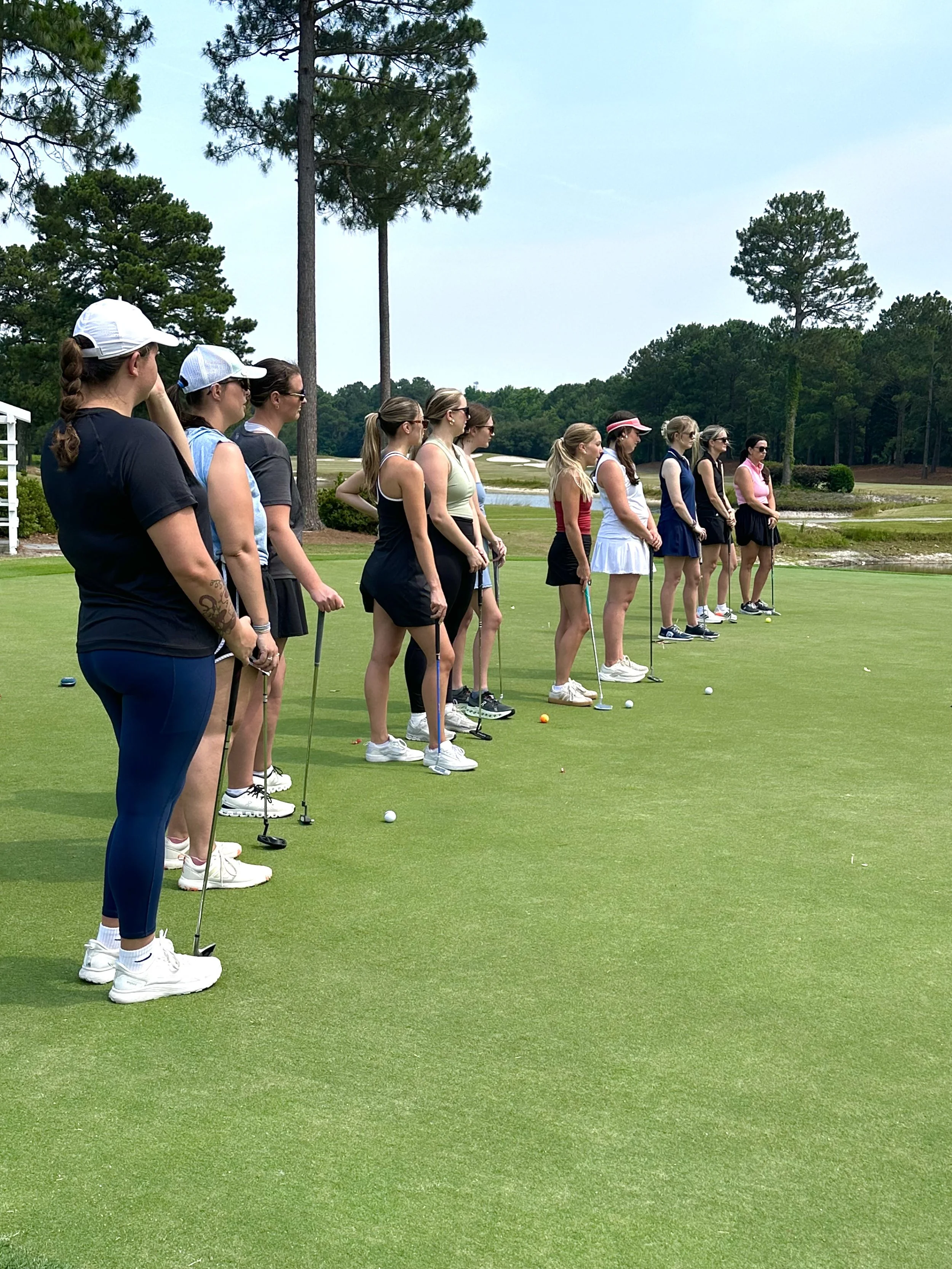 A line of women standing on a golf course greens, preparing for a putting practice or game, wearing athletic clothing and hats, with trees and a pond in the background.