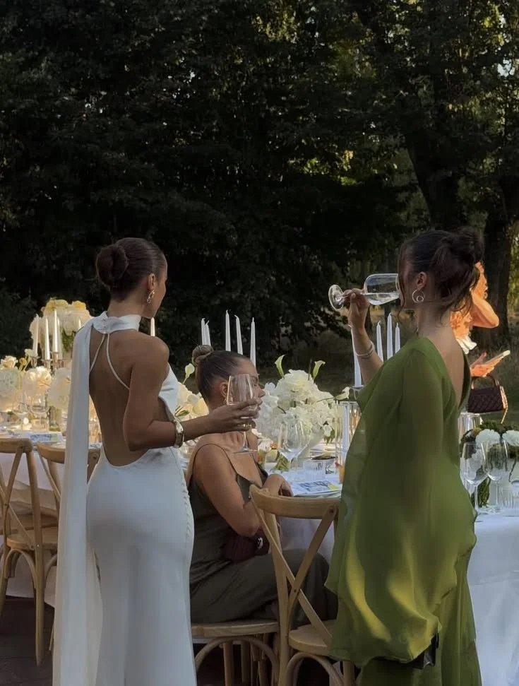 Three women at an outdoor dinner party, two standing and one sitting, with elegant table settings and floral arrangements in the background.