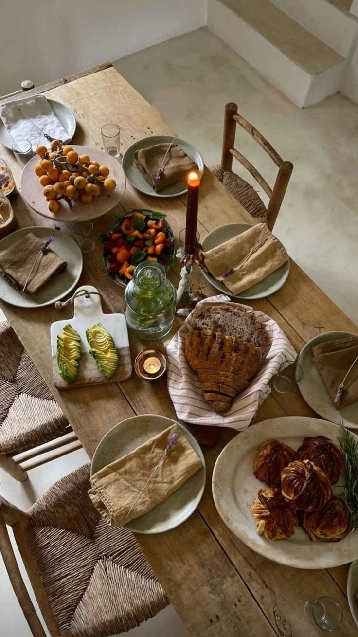 A rustic wooden dining table set for a meal with plates, cloth napkins, a bunch of apricots, a bowl of strawberries and other fruits, sliced avocado, sliced bread, croissants, a lit candle, and a glass jar with a green liquid.