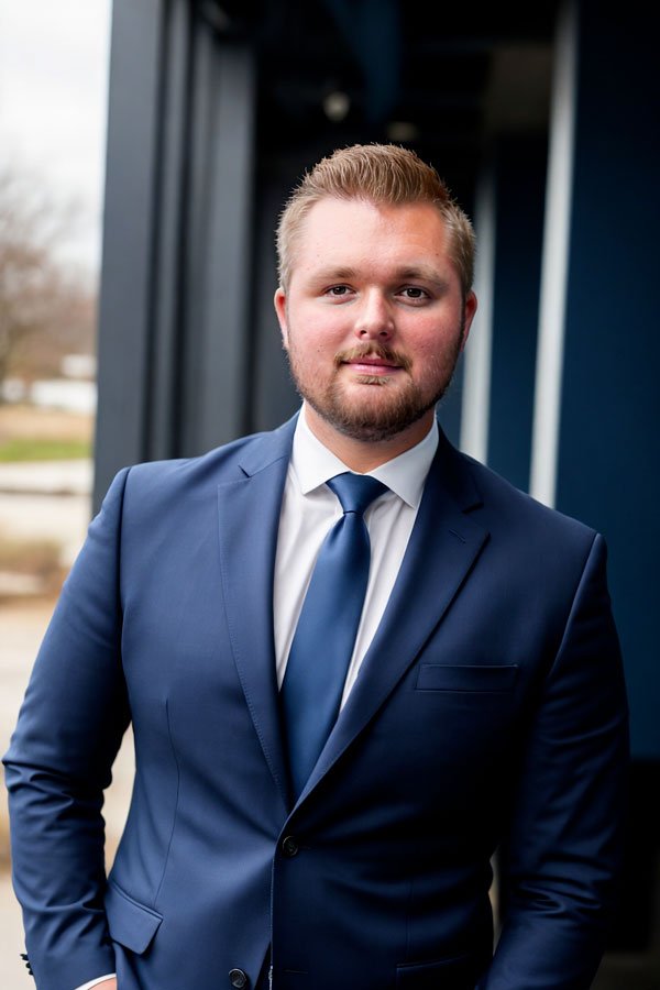 A young man with a beard and light brown hair, wearing a navy blue suit, white shirt, and a blue tie, standing outdoors in front of a building with blurred background.