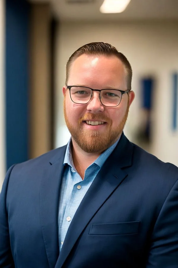 A smiling man with glasses and a beard, wearing a blue suit and light blue dress shirt, in an indoor setting.