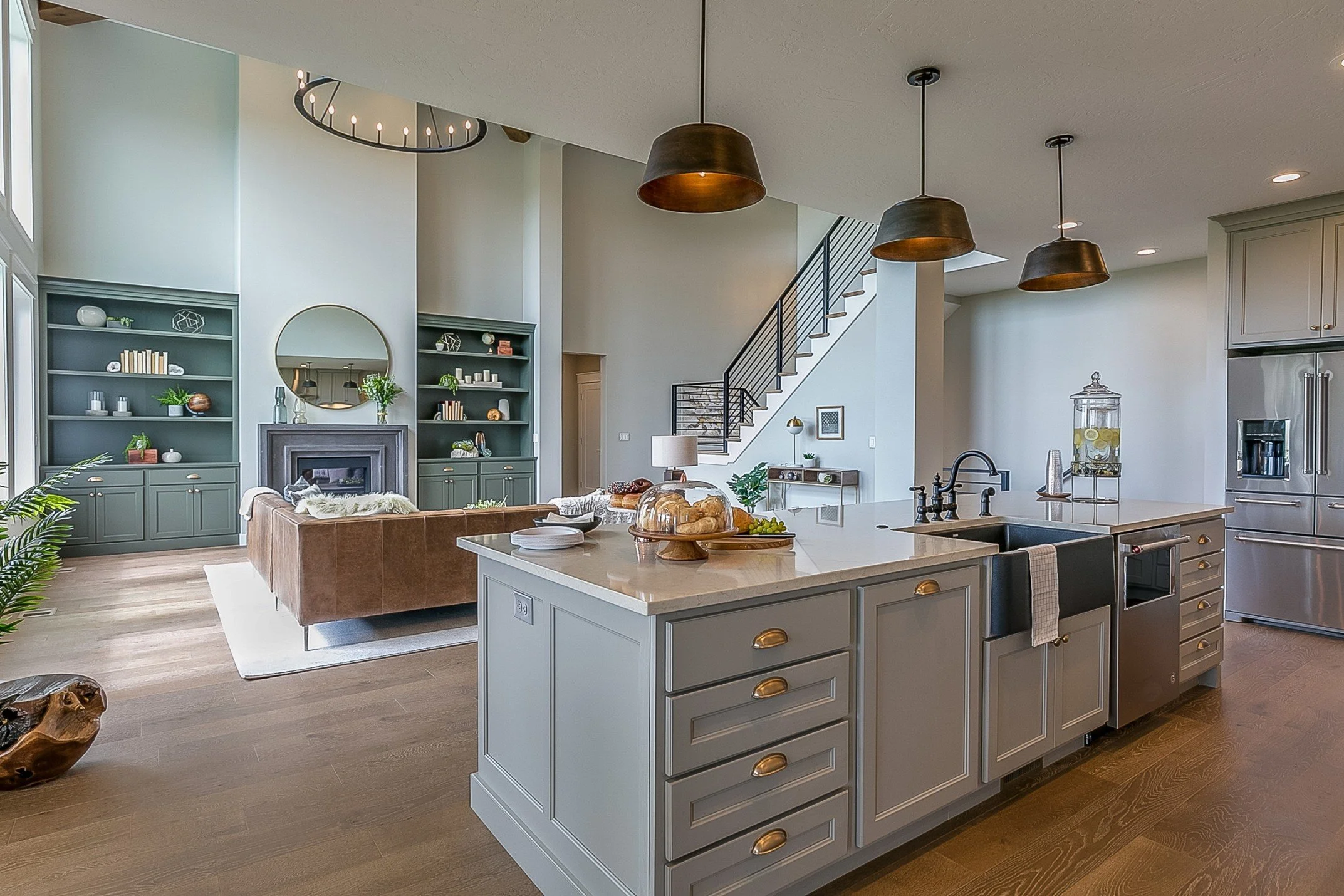 Modern open-concept kitchen and living room with high ceilings, pendant lighting, built-in shelves, a staircase, and a kitchen island with fresh bread, grapes, and snacks.