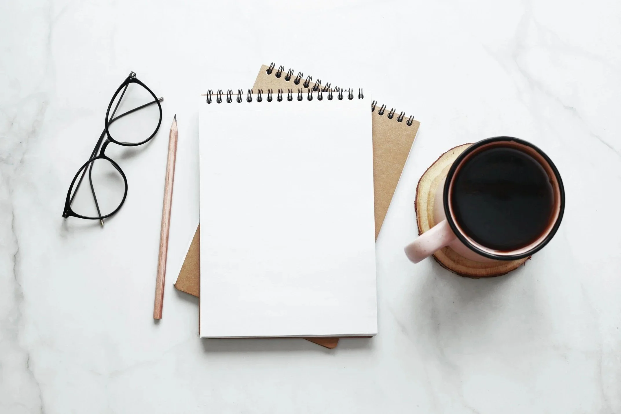 A flat lay of a workspace on a white marble surface, including black eyeglasses, a pink pencil, a spiral notebook, a brown notepad, a blank notepad, a coffee cup with black coffee on a wooden coaster.