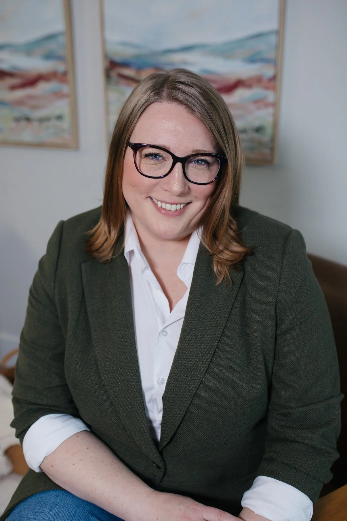 A woman with shoulder-length brown hair, wearing glasses, a white shirt, and a dark green blazer, smiling while sitting indoors. There are two abstract landscape paintings hanging on the wall in the background.