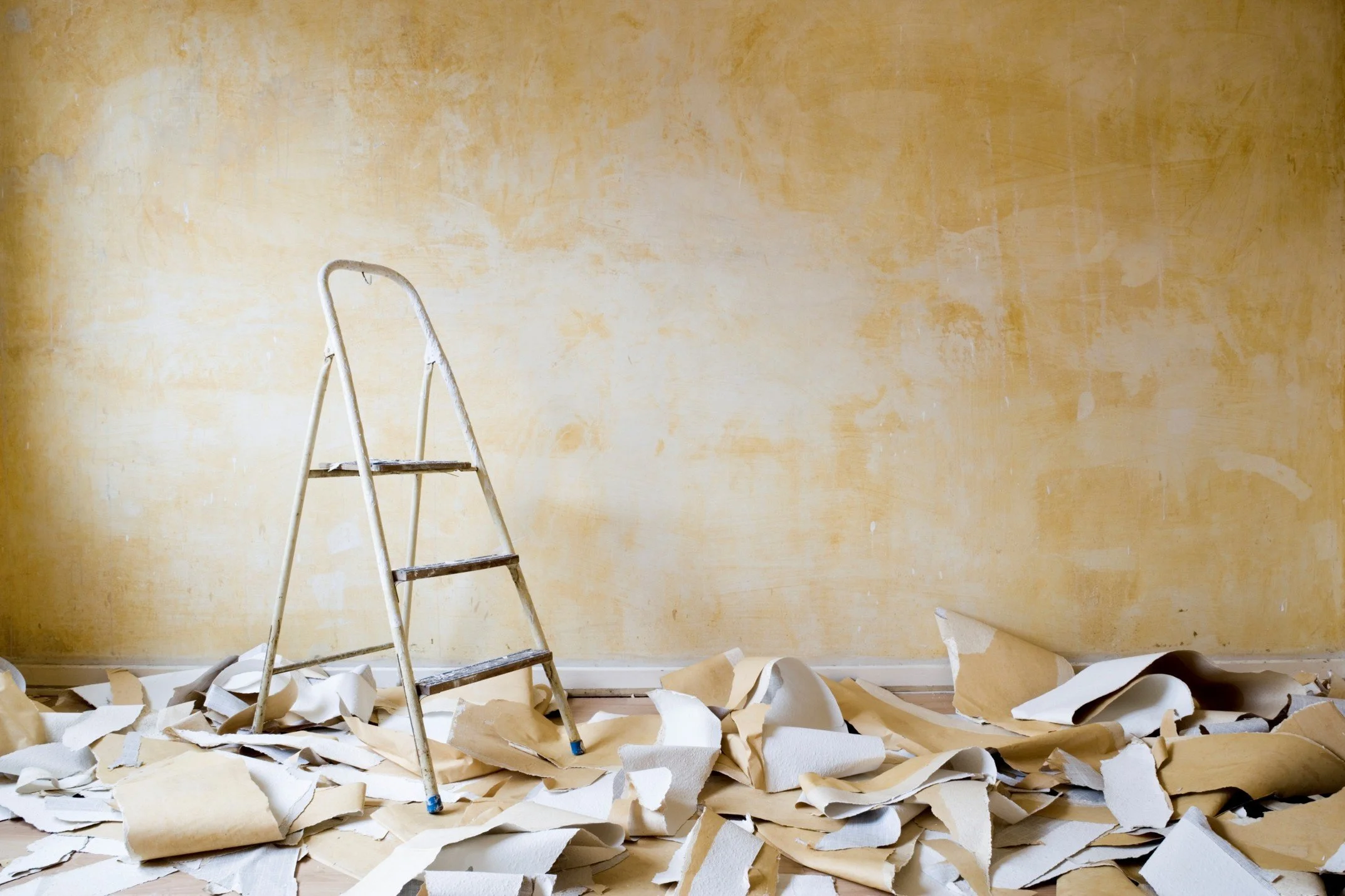 A rusty metal step ladder is in the center of a room with yellowing, unfinished wall and flooring covered with crumpled painter's paper sheets.