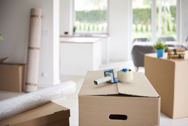 Open moving box with tape dispenser on top, situated in a bright living room with large windows and furniture.