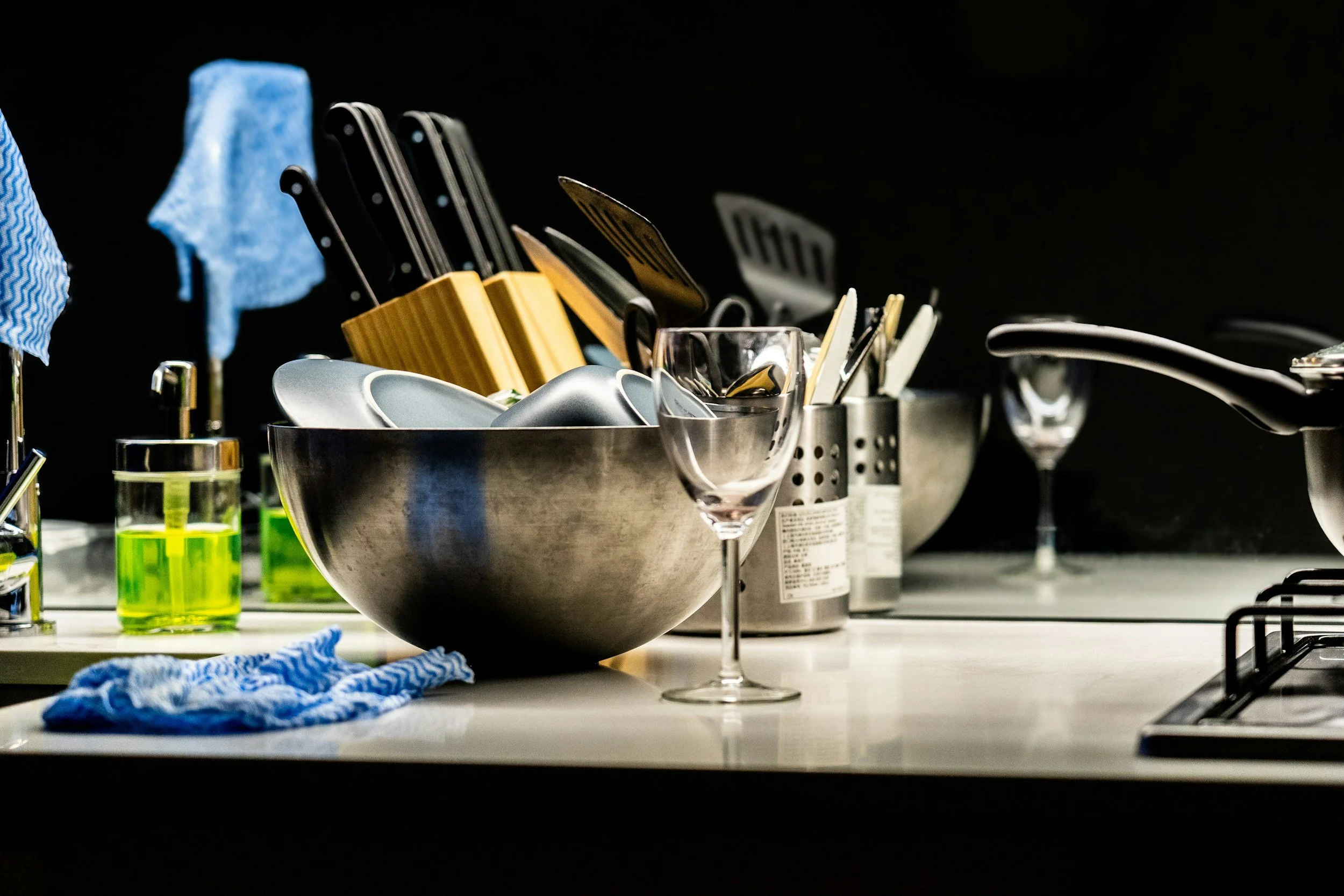 Kitchen countertop with a metal bowl holding cutlery, clear wine glasses, green liquid in glasses, yellow utensils holder, utensils, and some cleaning cloths. The background includes a stove and other kitchen items, all against a dark backdrop.