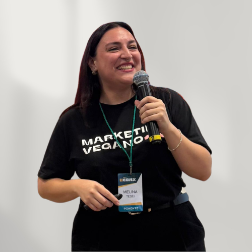 A woman with dark hair smiling and speaking into a microphone. She is wearing a black T-shirt with white text and a conference badge around her neck.