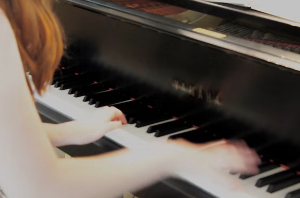 A young woman with red hair is shown close up playing a black grand piano.