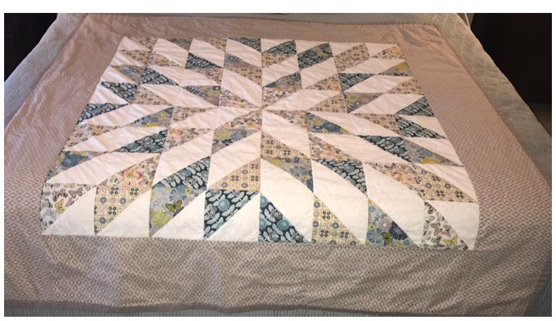 A quilt with a floral and geometric pattern in shades of blue, beige, and white, laid out on a bed.