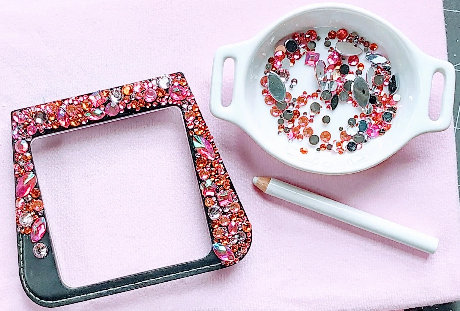 A picture of a pink surface with a decorated square picture frame covered in pink and clear rhinestones, a white ceramic bowl filled with various rhinestones and rhinestone settings, and a white stylus or pen.