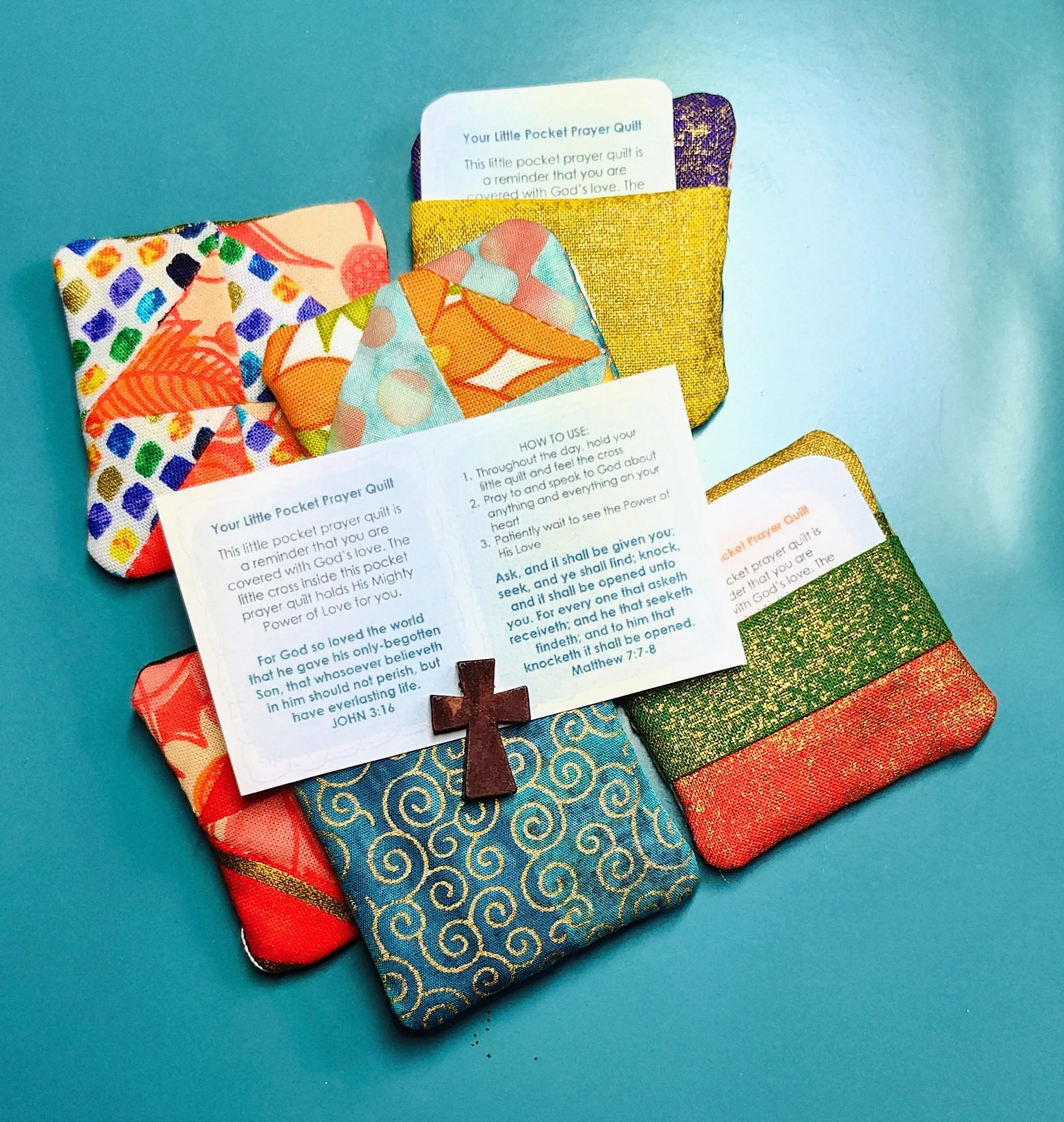 Colorful fabric pocket prayer quilts with a prayer card tucked inside, displayed on a teal background.