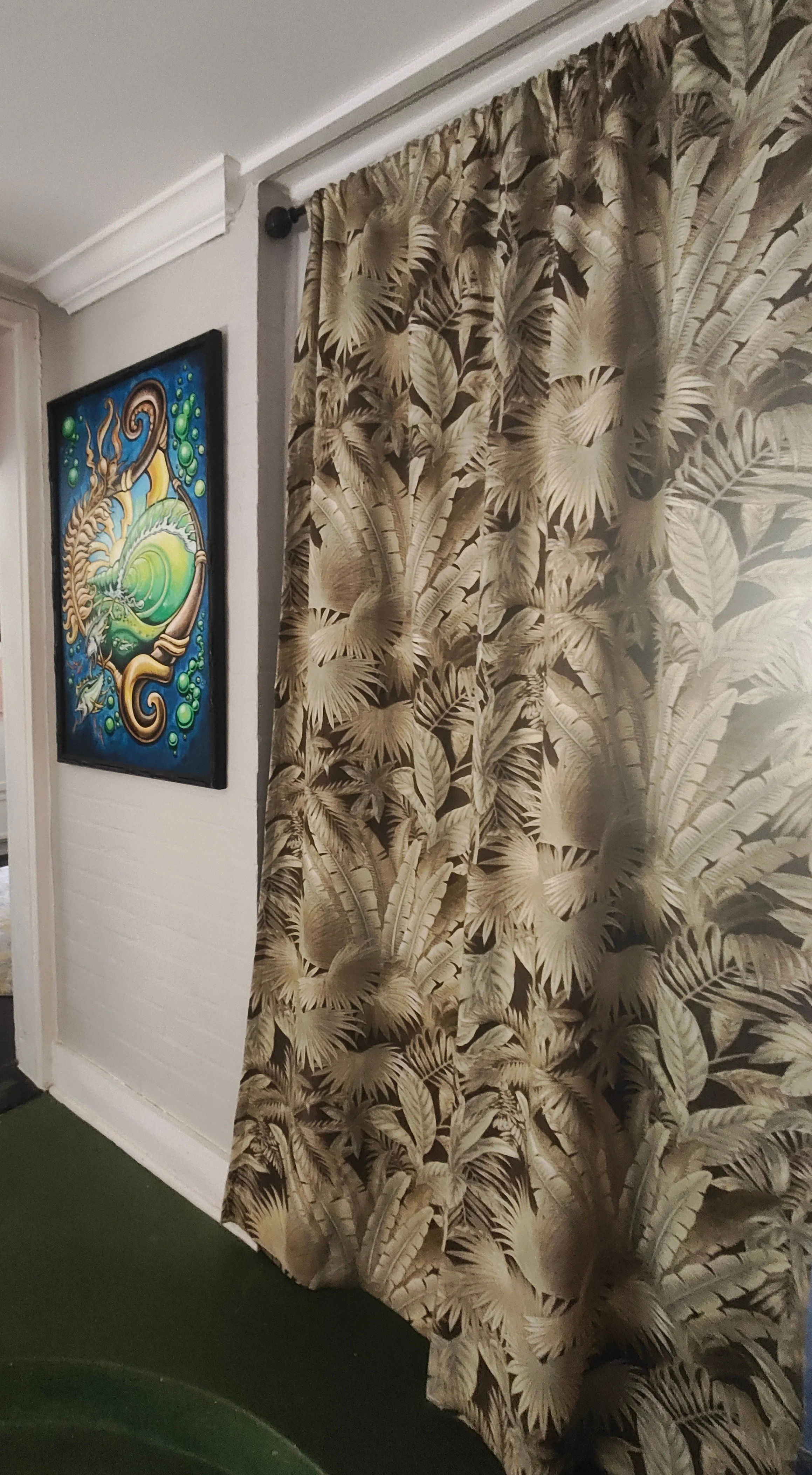 A hallway with tropical-themed curtains featuring large palm leaves, a colorful abstract painting on the wall, white walls, and green flooring.