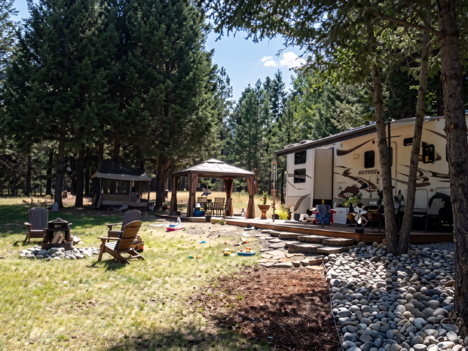 A campsite with a large trailer, two gazebos, wooden chairs, and a fire pit surrounded by rocks in a forested area.