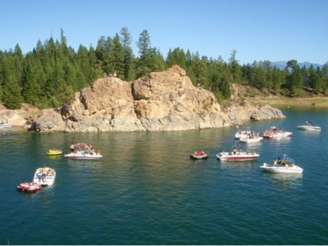 Boats anchored near a rocky shoreline with tall trees and hills in the background on a clear, sunny day.