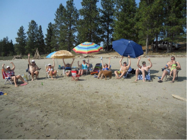 Madera Ranch Resort BC People relaxing on the beach under umbrellas, sitting on chairs, some with raised arms, with trees and parked boats in the background.