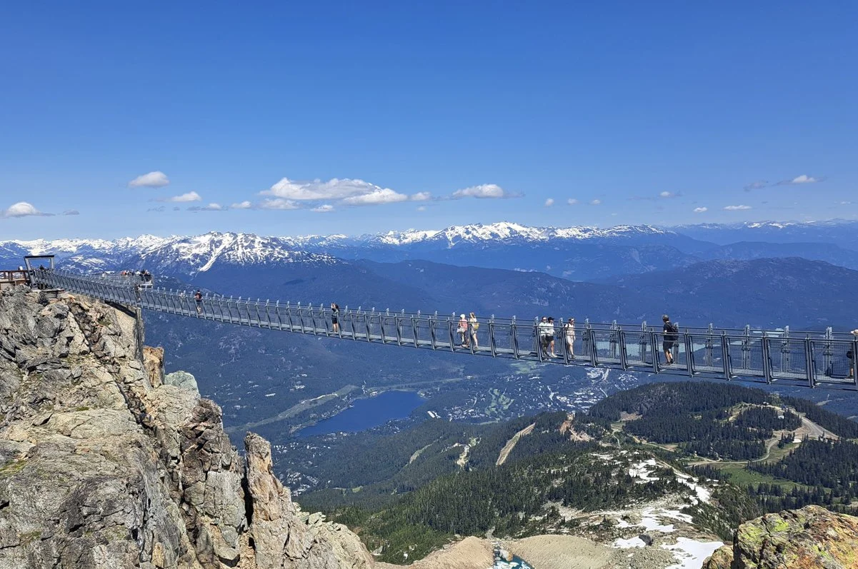 A suspension bridge on a mountain with people walking on it, overlooking snow-capped peaks and green valleys below on a clear day.