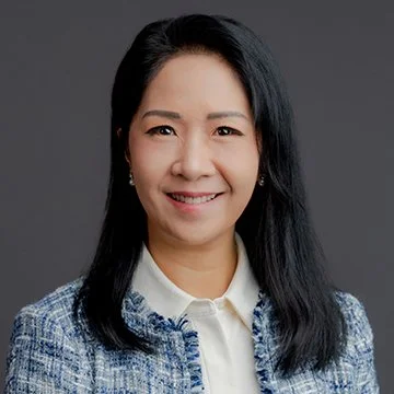 A woman with long black hair smiling, wearing a white collared shirt and a blue checkered blazer, against a gray background.