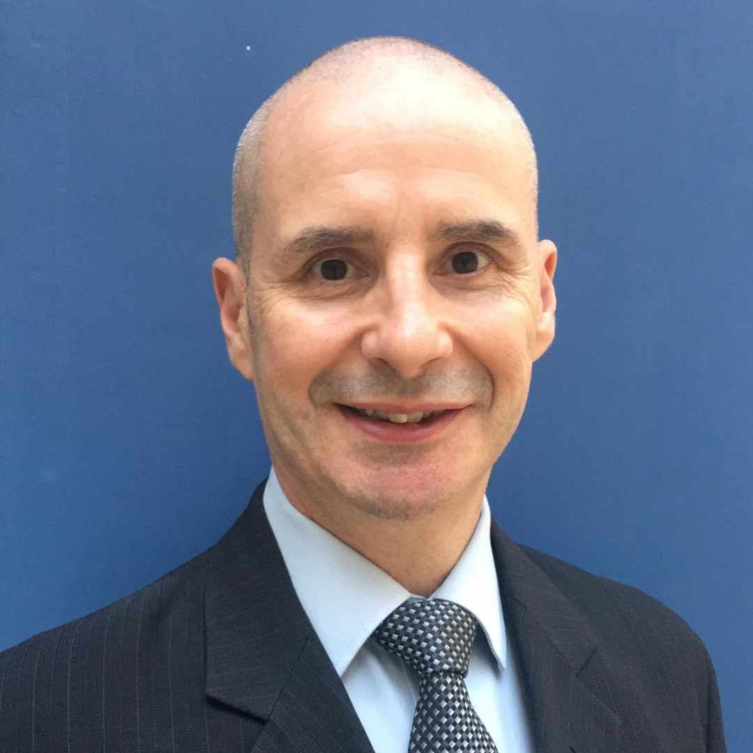 A middle-aged man with a bald head and light skin, wearing a dark pinstripe suit, a light blue shirt, and a patterned tie, smiling at the camera with a solid blue background.