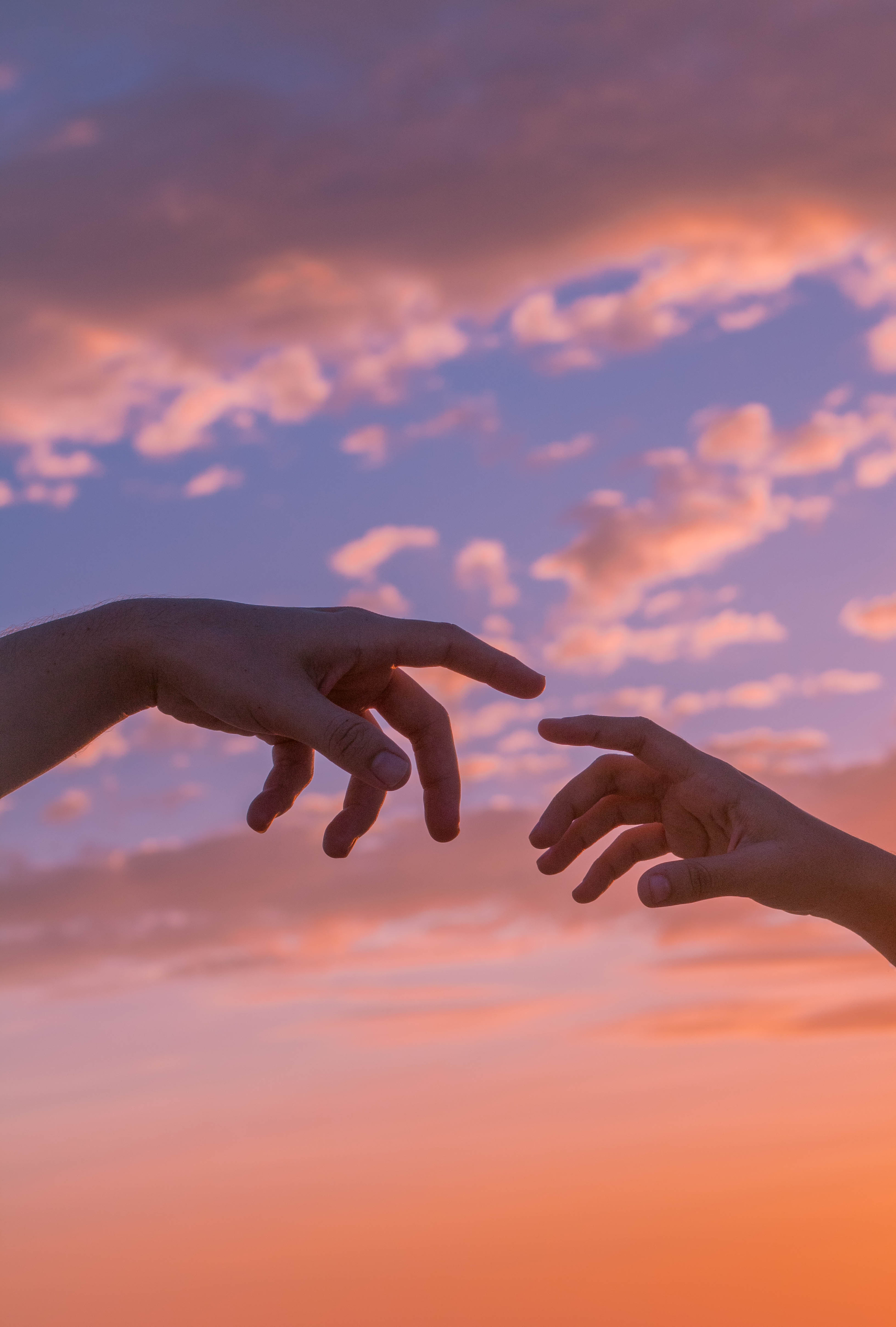 Two hands reaching towards each other against a colorful sunset sky with pink and purple clouds.