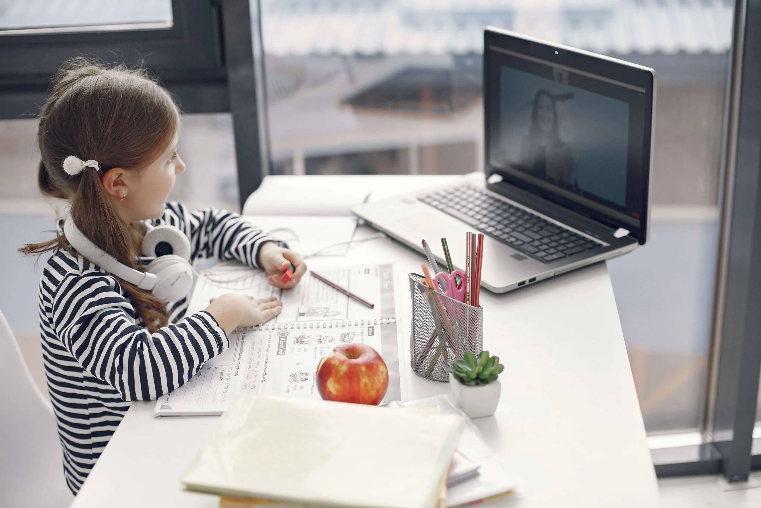 A young boy with headphones raising his hand and smiling at a laptop, with a lamp and window in the background.
