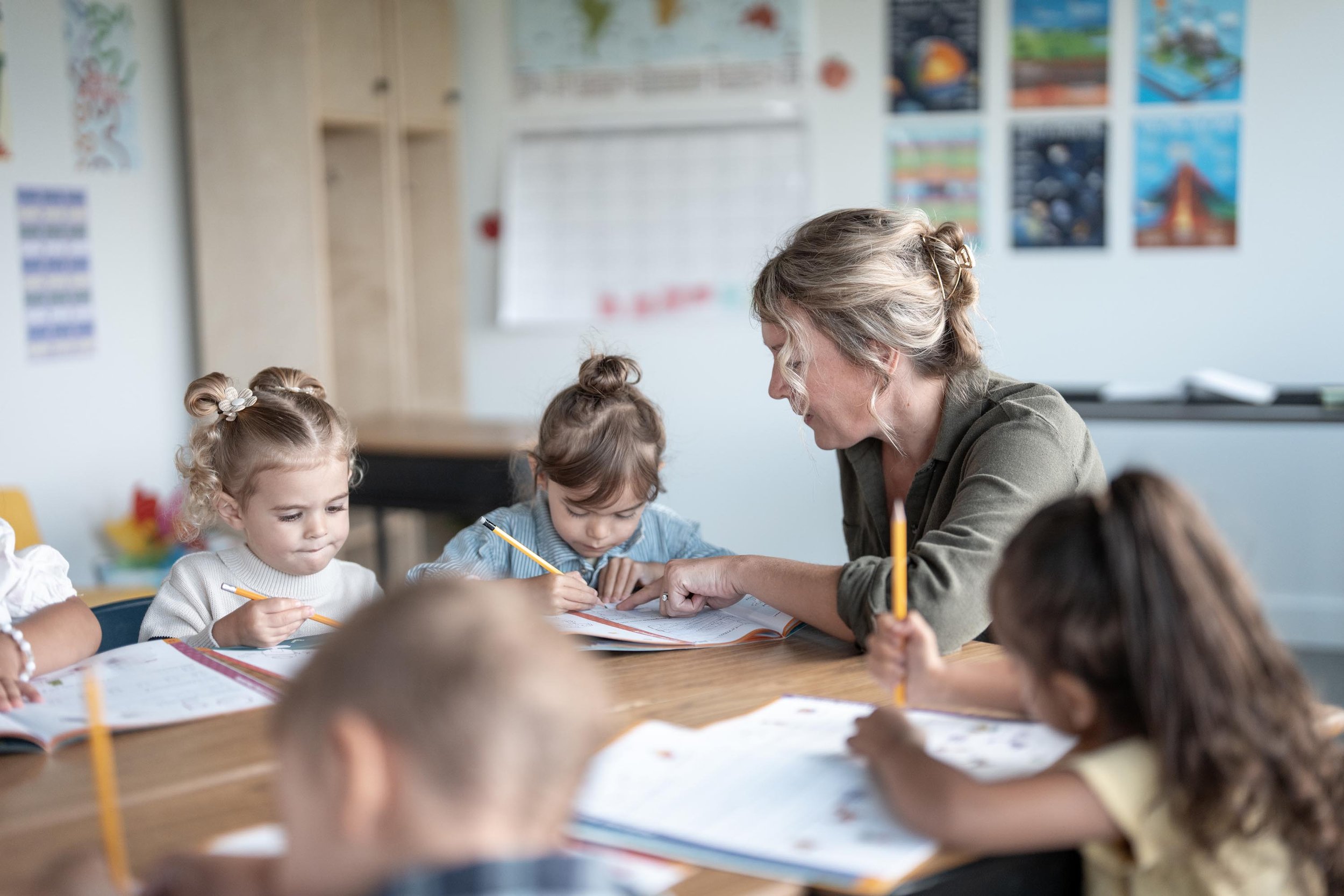 A classroom scene with a teacher engaging with young students at a table, all holding pencils and appearing focused on their work.